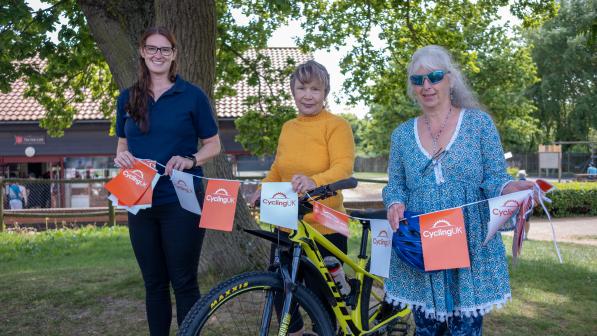 Three smiling women stand outdoors beside a yellow mountain bike decorated with Cycling UK-branded bunting. One wears a navy polo shirt, one a mustard yellow jumper, and one a turquoise patterned tunic with sunglasses. They are under a large tree with a red-roofed building in the background on a sunny day.