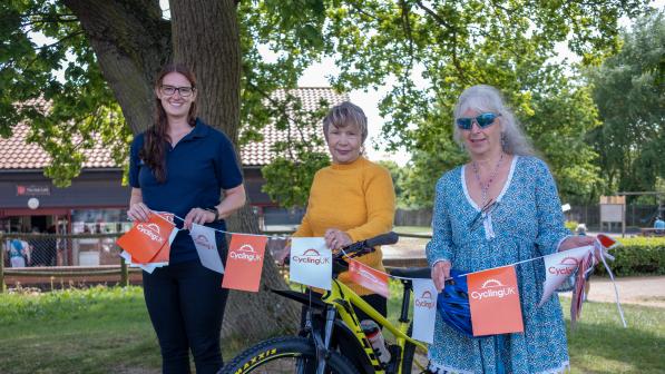 Three smiling women stand outdoors beside a yellow mountain bike decorated with Cycling UK-branded bunting. One wears a navy polo shirt, one a mustard yellow jumper, and one a turquoise patterned tunic with sunglasses. They are under a large tree with a red-roofed building in the background on a sunny day.