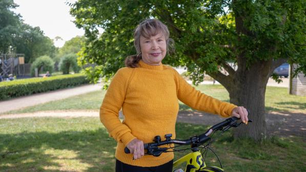 A woman wearing a yellow jumper stands beside a mountain bike in a sunlit park, smiling with one hand on the handlebars and a large tree in the background.