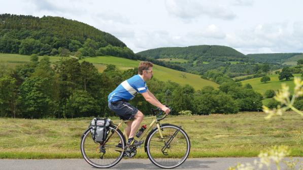 A man is riding a touring bike along a paved road with hills and forest in the background. He's wearing shorts and T-shirt