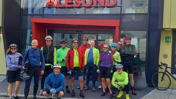 A group of people in cycling kit is standing in front of a hotel with a sign in red letters reading 'Alesund'. A black road bike is leaning against the wall of the hotel