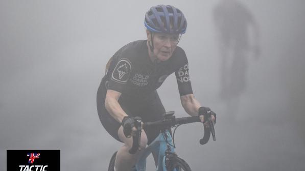 Deborah John riding a road bike in full cycling gear. She has a focused look on her face and is surrounded by a hazy mist, which makes the rider behind her hard to see, but gives the photo and eerie but beautiful quality.