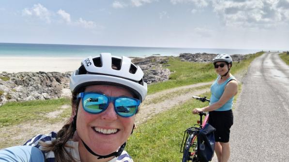 Diane Clayton has taken a selfie. She is wearing a white cycle helmet and cycling sunglasses. Another cyclist is behind her, and in the background is a dry stone wall and beyond that the sea.