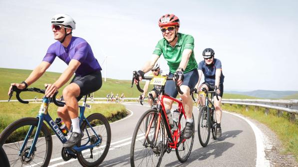 Four people in cycling kit are riding road bikes on a road going up and around a mountain. They have numbers on their bikes and helmets