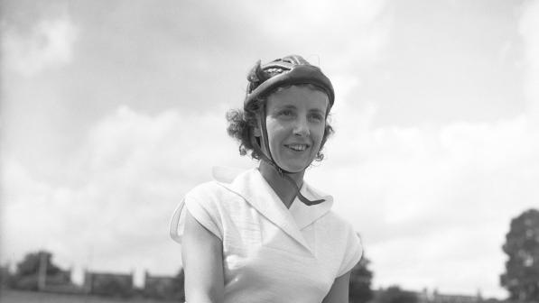 Eileen Sheridan is pictured in a black and white photo at Herne Hill cycle track in London. She is sitting on her Hercules bike wearing shorts and a short-sleeved top and cycle helmet and is smiling