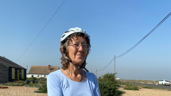 Eilidh Murray posing with her bike, wearing a helmet, light blue T-shirt and darker, smoky blue shorts. She is at Dungeness, in front of Prospect Cottage, the former house of artist Derek Jarman