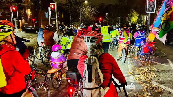 A big group of people is cycling along a city street at night. They're stopped at a red traffic light. There are adults and youngsters. There's leaves on the ground and the riders are wearing warm jackets