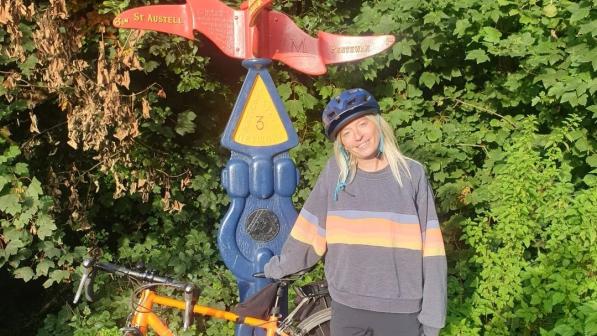 Photo of Ellie Lins posing by a cyclepath signpost with her orange bike. It's sunny, there's a forest in the background and Ellie is smiling, wearing a sweater and shorts.