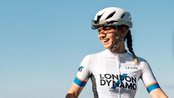 A photo of Ellie Cousins smiling. She is wearing a white helmet and a white short sleeved jersey with the London Dynamo logo, with blue sky behind her.