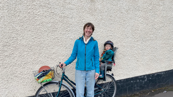 Emily Ryder is standing by a white wall with her cycle leaning up against the wall. She has an assortment of things in her front basket and her young child is sitting in the rear child seat