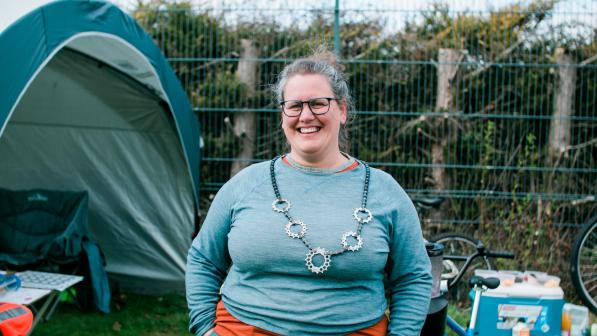 Emily Williams standing with her hands in her pockets, wearing a blue long-sleeved top, glasses and is smiling. She has a necklace made from a bike chain