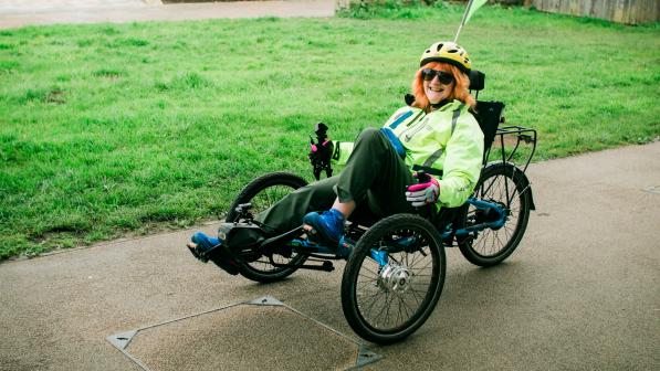 Smiling rider wearing a hi-vis jacket, helmet and sunglasses sits on a low recumbent tricycle with a safety flag, on a paved path beside a grassy area.