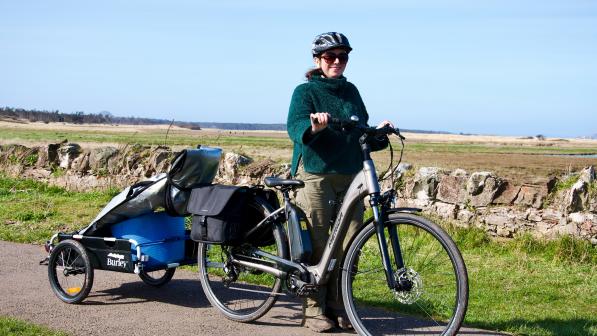 A woman in work clothes is standing holding an electric bike with a trailer attached and panniers on a cycle path with a dry stone wall and fields in the background