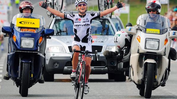 Emma Pooley cycling with her hands in the air in celebration as she finishes a race. She is wearing black and white racing gear, a helmet and cycling glasses, and has a wide grin. Safety cars and motorbikes are tailing her celebratory lap.