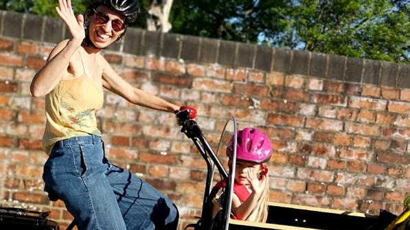 Emma Street waving at the camera and smiling while cycling. She's riding a cargo-bike with a child in it.