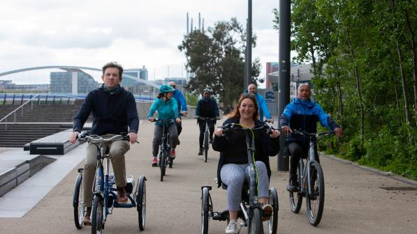 A group of people are cycling along a paved off-road path. They are on a mix of bikes but all are e-bikes.