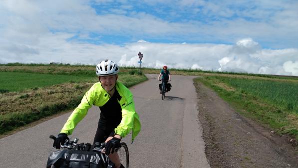 A youngster on flatbar bike with a packed bar bag is riding along a paved road in the countryside. They're wearing cycling kit and helmet and smiling at the camera. An adult on a bike is in the background