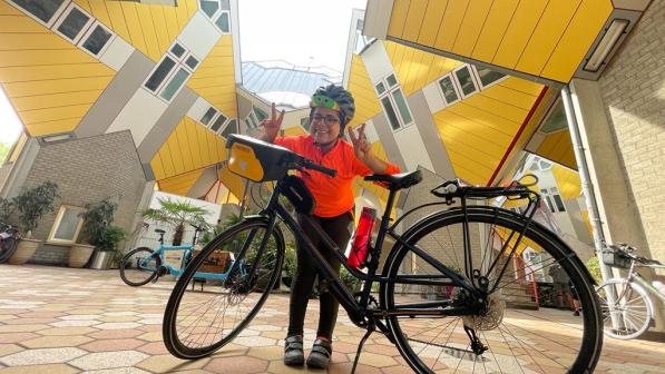 Fatema Abuidrees is standing behind a parked black bike smiling and making peace signs with her hands at the camera. There are surreal, diagonal yellow walls behind her in the background.