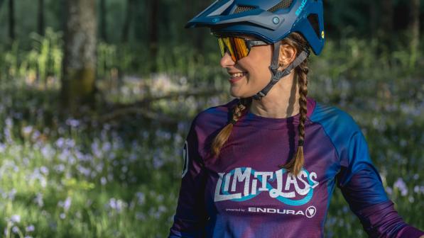 Fiona Finnie is standing in a sunny flower covered woodland wearing a cycle helmet, sunglasses and a t-shirt that reads Limitlass