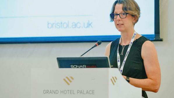 Fiona Spotswood behind a lectern with a laptop and mic propped on it. She is giving a lecture at a university.