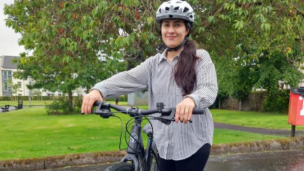 A woman is standing with her bike on a wet path in a park. She's wearing a shirt and leggings and a cycle helmet