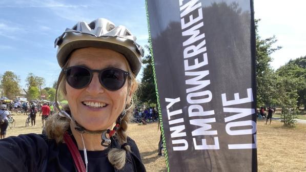 Gönül Ekmekçi Tekin is wearing sunglasses and a cycling helmet and is standing next to a black flag that reads ‘unity empowerment love’ there are crowds of people behind her some with cycles