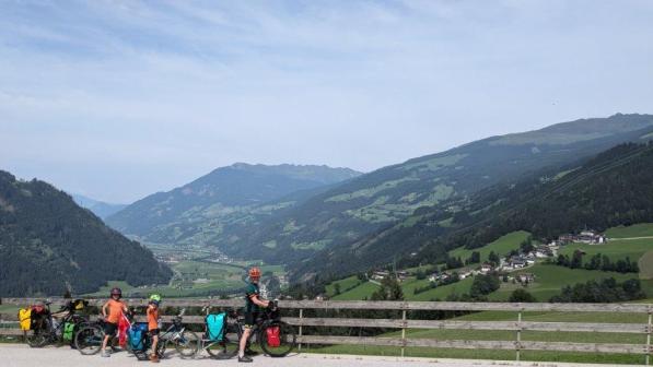 A man and two children are standing next to a fence with four bikes leaning on it. There are Alpine mountains in the background.