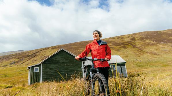 A woman in a red jacket and black trousers with a black headband is holding a mountain bike. There's a green hut in the background and grass-covered mountains
