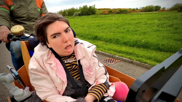 Greta Rafi McMillan is wearing a pink jacket sitting on the front of a cargo bike. There is a grassy hill in the background