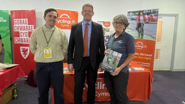 Three people are standing in front of a table in a conference room. The table is covered with an orange Cycling UK cloth and has Cycling UK posters behind it