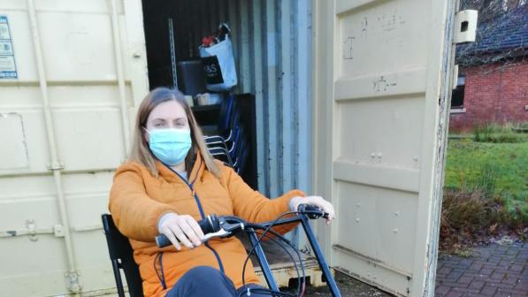 Hannah Knox is sitting on a recumbent tricycle in front of a white shipping container she is wearing a blue face mask and yellow jacket