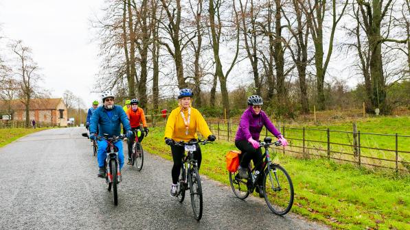 A group of cyclists ride along a quiet country lane lined with bare trees in winter. The riders are dressed in bright jackets and helmets, with the lead woman in yellow smiling confidently. The road is wet and bordered by grassy verges and wooden fencing, suggesting a cool, damp day in the countryside.