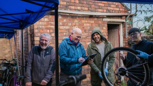 Four men smile and chat while repairing bikes outdoors under a canopy, with one holding a clipboard and another holding up a bike wheel.