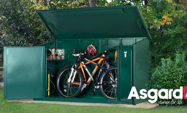 Two mountain bikes and a floor pump are in a green Asgard bike storage shed in a garden. The Asgard logo is on the bottom right of the photo
