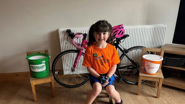 Heidi Barker is sitting on a chair in front of her pink bike. On either side of her bike are charity donation buckets and she is wearing an orange t-shirt and cycling shorts