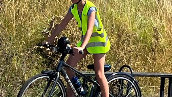 A young cyclist wearing a high-visibility vest, sunglasses, and a helmet is riding a bike along a sunny rural path surrounded by tall grass.