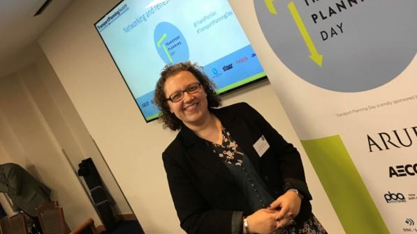 Helen Hemstock is wearing a black blazer jacket and floral dress and is standing in front of a roller banner that has the words 'Transport Planning Day' at the top