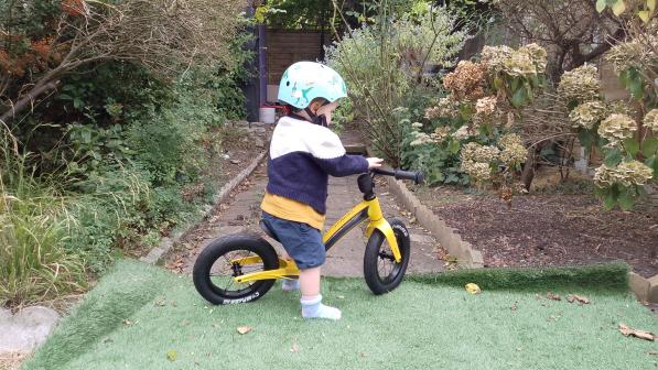 A little boy is riding the Hornit AIRO Balance Bike, a yellow balance bike. He's wearing a helmet and he's in a garden