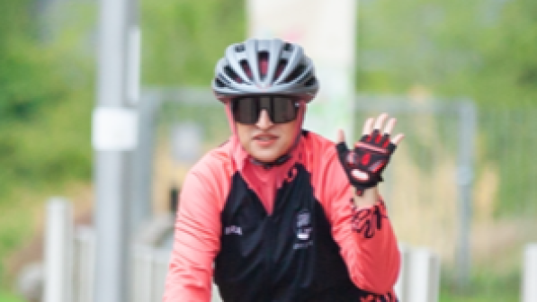 Humira Shahzad is cycling along a wide cycle path, wearing a red top, sunglasses and black cycle helmet. She is waving with one hand