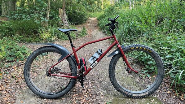 A red mountain bike with wide tyres, flat bar and mudguards fitted is propped up on a muddy track in a forest