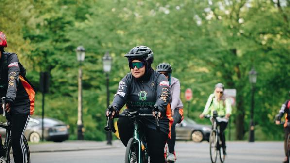 Iffat Tejani is cycling along a road with several other women. She is wearing sunglasses and a cycle helmet with a head scarf underneath