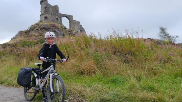 Action shot of Iona O’Donnell on her bike in front of castle ruins, nominated as a cycle influencer in Cycling UK’s 100 Women in Cycling 2024