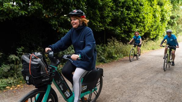 A woman riding an e-bike on a tree-lined gravel path, smiling and wearing a helmet and a navy jacket. Behind her, two other cyclists ride traditional bikes, all enjoying the outdoor setting.