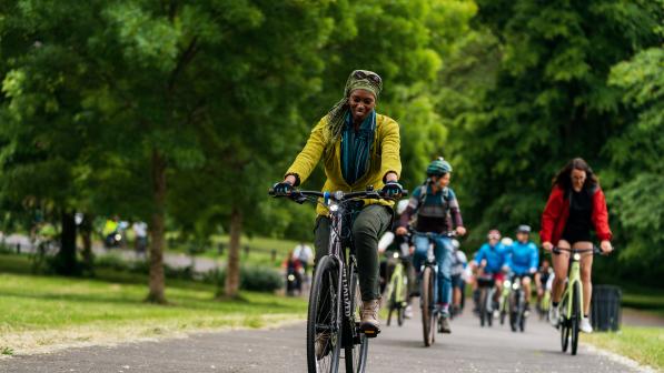 A large group of people is cycling along a paved path through green space, with trees behind them. There is a woman cycling on an e-bike at the front. She is wearing normal clothes and a headscarf. She is smiling