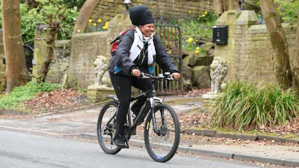 A woman in black jeans, jacket and turban is cycling along a road on a black mountain bike. She's smiling