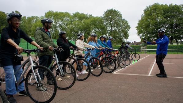 A cycling instructor in a blue jacket gives directions to a diverse group of adults lined up on bicycles during a training session on an outdoor court. All participants are wearing helmets and appear attentive, with trees and fencing in the background.