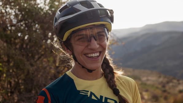 Jasmin Patel is pictured smiling wearing a cycle helmet and glasses with a mountain range and trees in the background