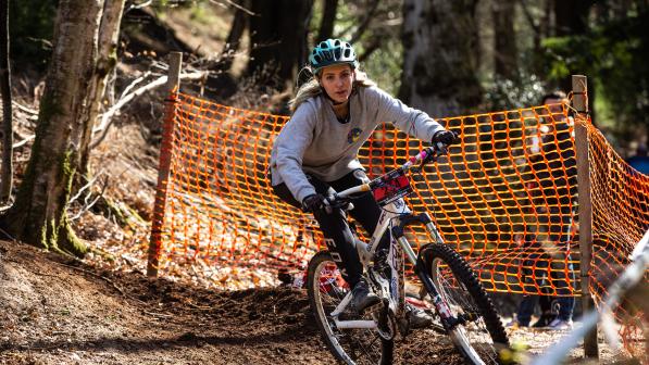 Jaz Morse is riding down a muddy woodland track on a mountain bike, there is an orange plastic fence and trees in the background