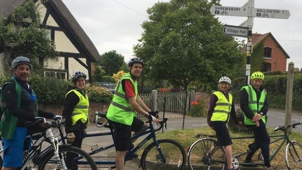 Jean Breakell is sat in the stoker position (at the back) on the tandem under a street signpost in a village. There are other cyclists around her, all wearing high vis vests