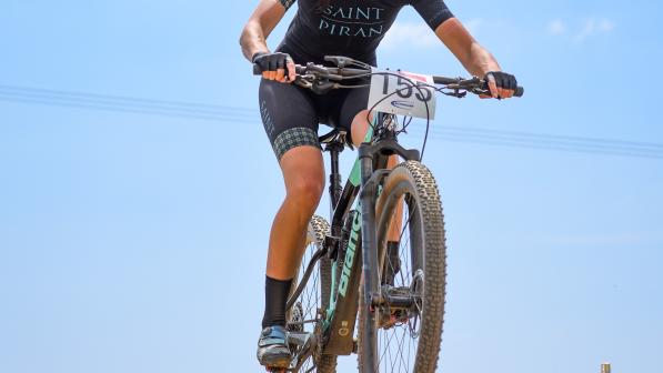 Jenny Bolsom on her mountain bike wearing Saint Piran kit, a helmet and sunglasses. In the photo, her bike is up in the air above the gravel track below as she has just ridden over a bump.
